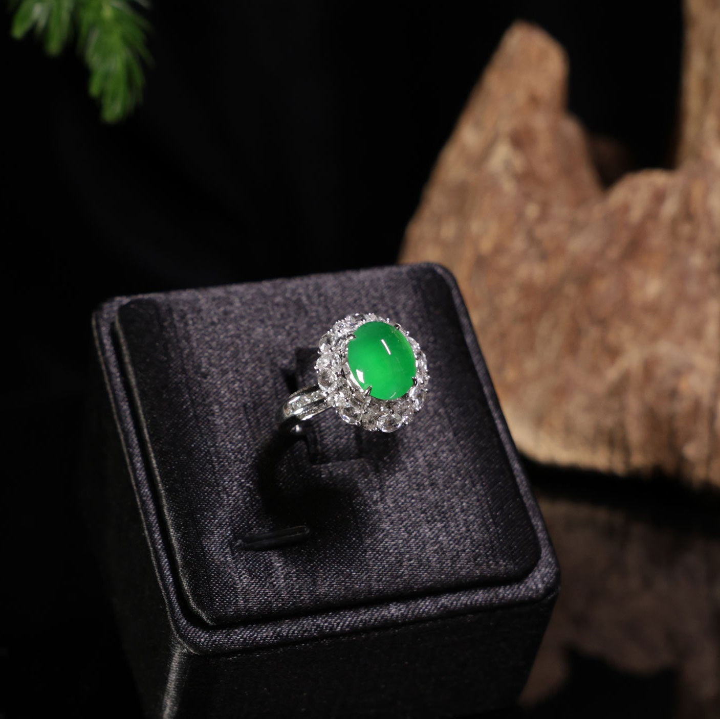 A halo ring featuring a round green jadeite stone surrounded by small clear stones, displayed on a black velvet jewelry box against a dark background with a piece of wood.